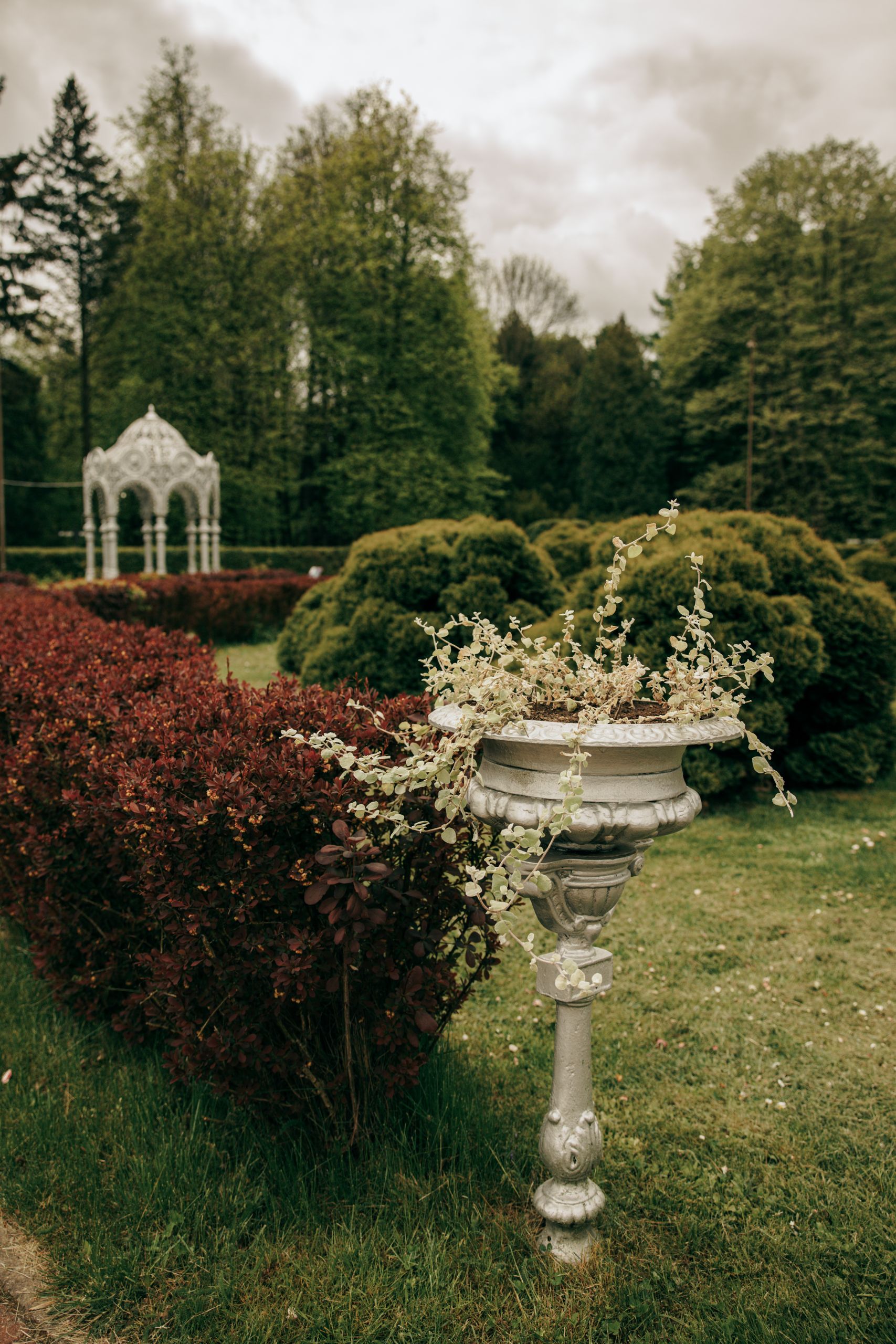 A picturesque garden with a vintage vase, lush greenery, and a distant gazebo under cloudy skies.